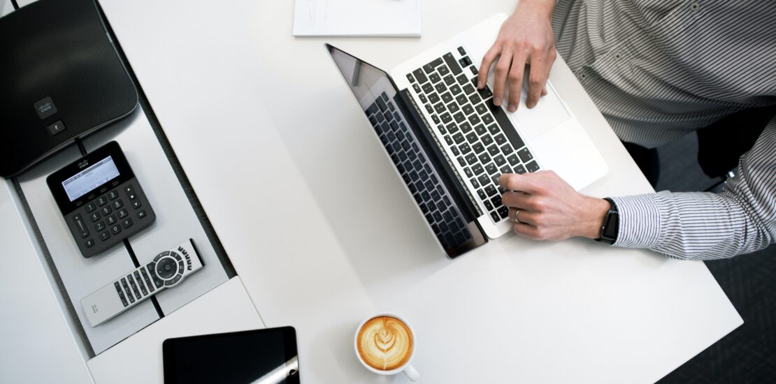 homem sentado em uma mesa mexendo em um notebook com outros aparelhos e um café. SPED publica novas versões dos programas da ECD e da ECF