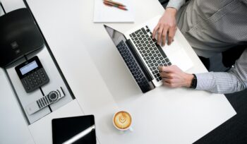 homem sentado em uma mesa mexendo em um notebook com outros aparelhos e um café. SPED publica novas versões dos programas da ECD e da ECF